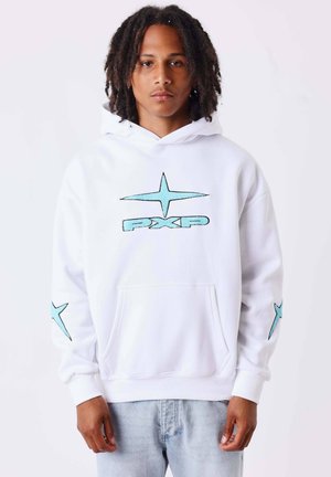 Jeune personne aux cheveux bouclés mi-longs portant un sweat à capuche blanc avec une étoile bleue et le logo "PXP", debout devant un fond uni.