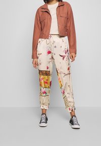 Avskuren rostfärgad jacka, beige topp och blommönstrade byxor med lapptäckesdetaljer. Svarta sneakers fulländar outfiten.