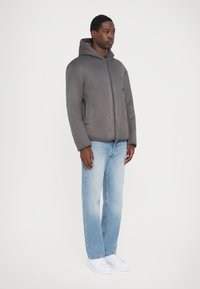 Grå huva jacka gjord av mjukt material, med en framtida dragkedja och sidofickor, bärs med ljusblå jeans och vita sneakers.