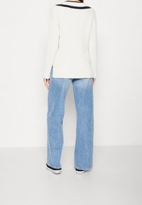 Person klädd i en vit ribbad tröja med svarta kragaränder, ljusblå vida jeans och svartvita Converse-sneakers, sedd bakifrån.