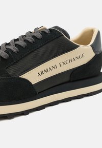 Tenisii din suede negru și plasă cu accente bej, având șireturi gri și logo-ul "Armani Exchange" pe lateral. Talpa din cauciuc texturat.