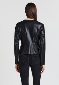 Donna con lunghi capelli castani che indossa una giacca di pelle nera attillata e jeans scuri, in piedi con le spalle rivolte verso la macchina fotografica su uno sfondo semplice.