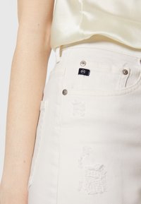 Vaqueros de denim blancos con una textura desgastada, herrajes plateados y una etiqueta de logo negra en el bolsillo delantero. Diseño ajustado visible.