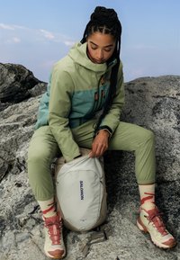 Giacca verde chiaro e teal con zip arancioni, pantaloni oliva coordinati, zaino beige con logo blu e scarpe da trekking rosa con accenti crema.