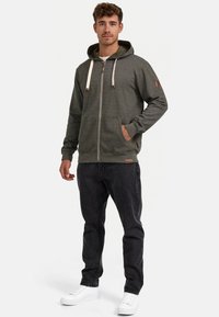 Sudadera verde con cremallera, capucha, cordones y bolsillos frontales. Combinada con pantalones negros y zapatillas blancas, con una textura sutil.
