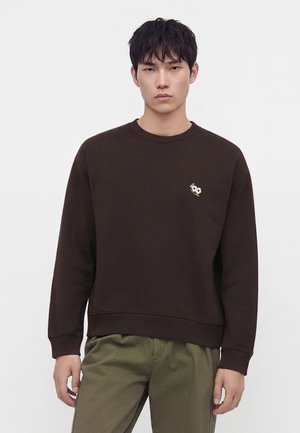 Jeune homme portant un sweat-shirt marron foncé avec un petit emblème blanc en forme de fleur sur la poitrine et un pantalon vert olive sur un fond uni.