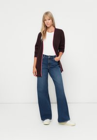 Cardigan long jusqu'aux genoux de couleur bordeaux foncé avec des bordures roses, associé à un jean bleu à taille haute et jambes larges et des baskets blanches. Texture lisse.