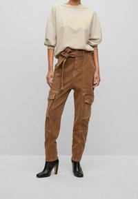 Beige collegepaita, jossa teksti "LONDON", yhdistetty korkeavyötäröisiin ruskeisiin cargo-housuihin, joissa on vyö, taskut ja sileä kangaspinta. Mustat korolliset saappaat.