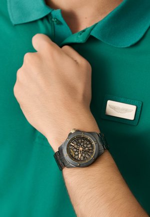 Orologio con quadrante a scheletro, cassa e cinturino in metallo. Presenta dettagli in tono dorato, quadrante rotondo e superficie testurizzata. Indossa una polo teal.