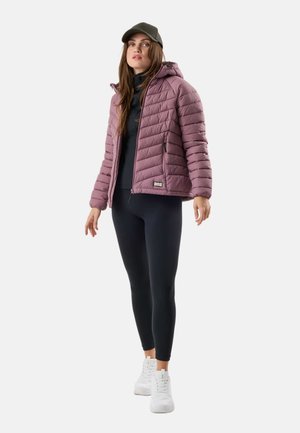Femme portant une veste matelassée mauve, des leggings noirs, un haut noir, des baskets blanches et une casquette de baseball vert olive, debout devant un fond blanc.