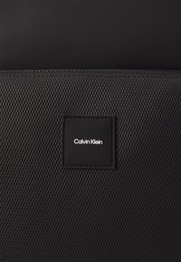 Zaino in rete nera con un'etichetta rettangolare del logo Calvin Klein, dettagli lisci e un design strutturato. Superficie testurizzata con un fine motivo a griglia.