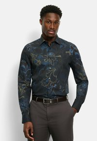 Schwarzes Hemd mit floralem Muster in Blau- und Grüntönen, ausgestattet mit Knöpfen und tailliertem Schnitt. Kombiniert mit dunkelbraunen Hosen.