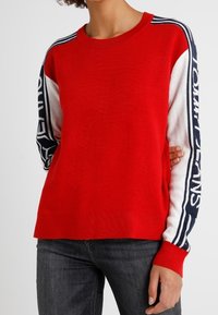 Czerwony sweter z białymi i granatowymi rękawami. Posiada okrągły dekolt oraz logo na rękawach. Wykonany z dzianiny.