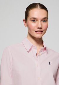 Light pink collared shirt with button front, featuring a small dark blue logo on the left chest. Smooth texture and tailored fit.