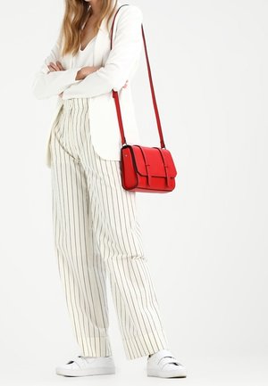 Vrouw in een wit colbert, witte gestreepte broek en witte sneakers staat met gekruiste armen, met een rode schoudertas.