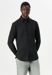 Svart langermet skjorte med knappeåpning og en enkelt brystlomme, med en glatt tekstur og strukturert passform, paired med lyse jeans.
