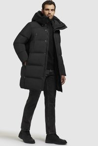 Cappotto imbottito nero con cappuccio, dotato di zip frontale, bottoni a pressione e tasche laterali. Indossato con jeans scuri e stivaletti neri.