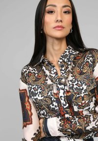 Chemisier à manches longues en tissu de soie à motifs, avec un riche design paisley en noir, or et rouge. Poches poitrine avec fermetures à bouton.