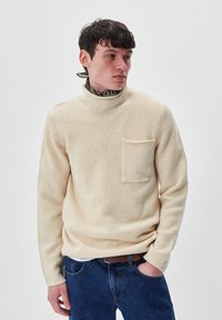 Beige gestrickter Pullover mit hohem Kragen, ausgestattet mit einer Brusttasche und gerippten Bündchen, kombiniert mit blauen Jeans. Strukturierter Stoff.