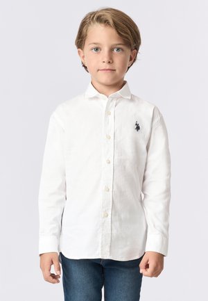 Camicia bianca a maniche lunghe con bottoni sul davanti, colletto e logo ricamato sul lato sinistro del petto. Abbinata a jeans blu in denim.