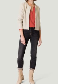 Veste en tissu crème, blouse rouge, jeans foncés avec les poignets retroussés, et bottines beiges. La veste n'a pas de boutons et dispose de poches avant.