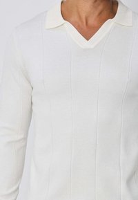 Personne portant un polo blanc à manches longues avec un col en V, montrant le haut du torse et la zone du cou.