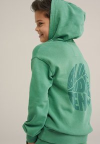 Sudadera verde con un bolsillo canguro en la parte delantera, tela texturizada y un gran gráfico impreso en la parte posterior. Cuenta con capucha ajustable con cordón.