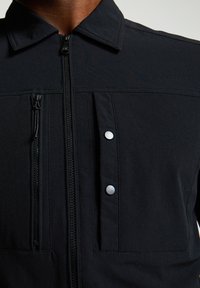 Melnā jaka ar rāvējslēdzēju priekšpusē, diviem sudraba pogām un rāvējslēdzēja kabatu kreisajā pusē. Izgatavota no gludas, strukturētas auduma.