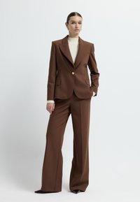 Bruine getailleerde blazer met een gesneden kraag, enkele gouden knop en twee zijzakken, gecombineerd met bijpassende wijde broek. Textured witte top eronder.