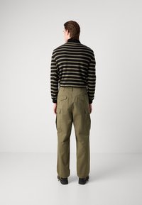 Gestreifter schwarzer und olivfarbener Pullover, lockere Passform, kombiniert mit olivfarbenen Cargo-Hosen mit Klappen-Taschen vorne und geradem Schnitt. Schwarze Schuhe.