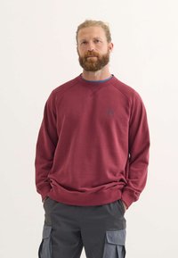 Felpa bordeaux con collo rotondo, polsini a coste e un piccolo logo sul petto. Abbinata a pantaloni cargo grigi. Texture liscia.