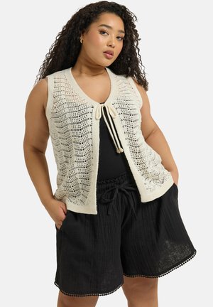 Strikket ærmeløs cardigan i creme med åbent vævemønster, lukning med bindebånd foran, parret med sorte, teksturerede shorts med bølget kant.