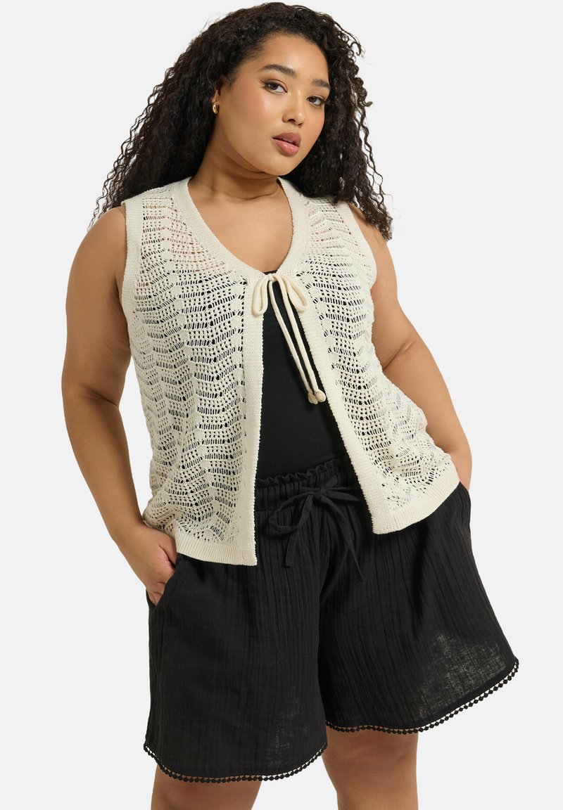 Stickad ärmlös kofta i krämfärgad nyans med öppet vävmönster, knytning framtill, ihop med svarta, texturerade shorts med scalloped fåll.