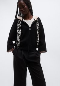 Sudadera negra con cremallera y detalles de estampado de leopardo en la capucha y la cremallera. Presenta un corte holgado y tela ligera.