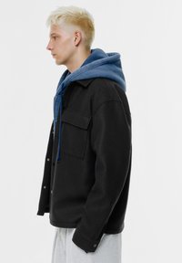 Černá bunda z vlněné směsi s volným střihem, náprsní kapsou a zapínáním na knoflíky, vrstvená přes modrou mikinu s kapucí a stahovací šňůrkou.
