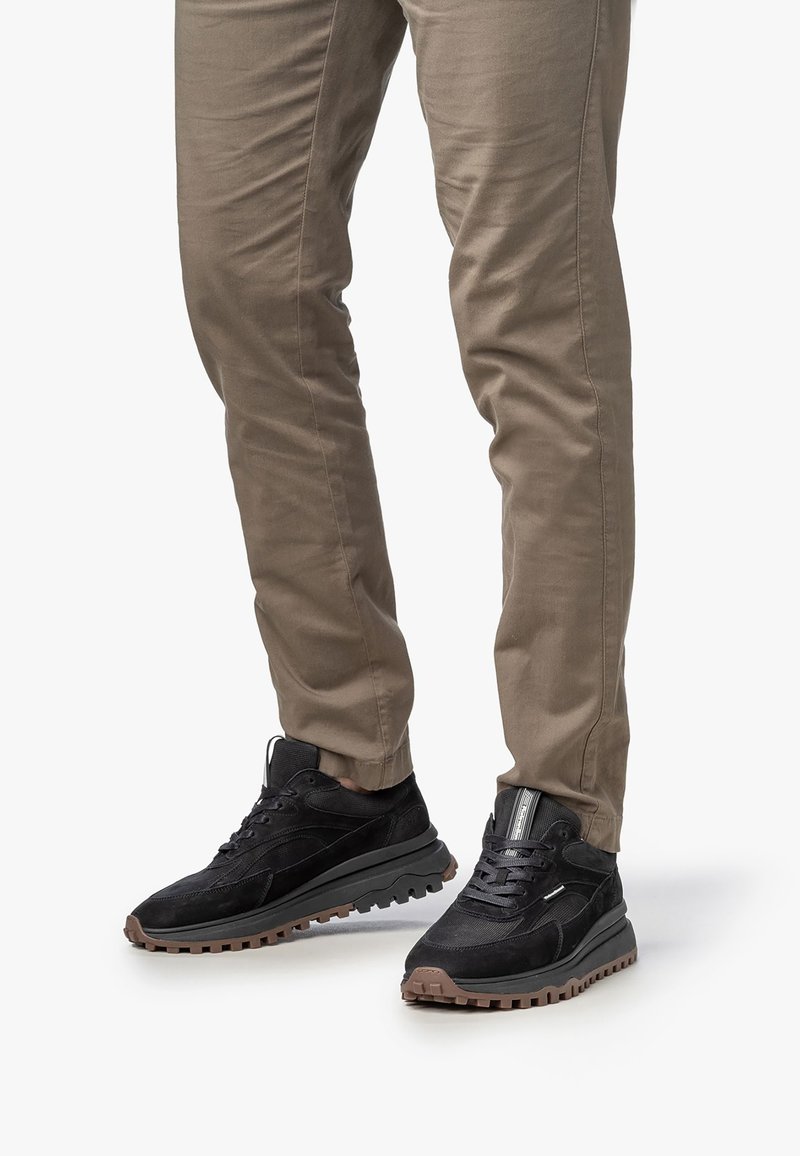 Svarta mockaskor med texturerade detaljer, gummisula och snörningsdesign, i kombination med ljusbeiga byxor.