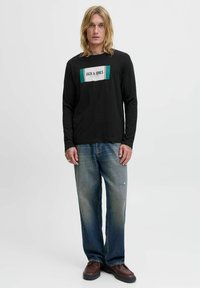 Schwarzes Langarmshirt mit einem rechteckigen weißen und grünen Logo. Blaue ausgewaschene Weitbein-Jeans und braune Schuhe runden das Outfit ab.