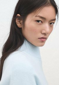 Svetlomodri rebrasti pulover z visoko ovratnico in mehko teksturo, ki ga nosi modelka z dolgimi temnimi lasmi, postavljena na nevtralno ozadje.