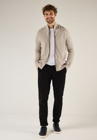 Cárdigan de cremallera beige claro con textura acanalada, detalles en cremallera negra, usado sobre una camiseta blanca, combinado con pantalones negros y zapatillas de deporte azul marino.