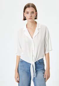 Camicia bianca con maniche corte, colletto e parte anteriore annodata. Realizzata in materiale leggero con una texture delicata. Abbinata a jeans blu.