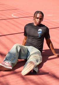 Schwarzes Nike-T-Shirt mit einem blauen Logo, kombiniert mit hellblauen Denim-Jeans und rosa Sportschuhen auf einem roten Platz.