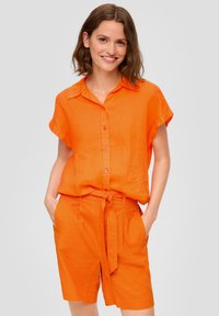Oranžová lněná košile s krátkým rukávem, límečkem a knoflíky, v kombinaci s odpovídajícími oranžovými šortkami s opaskem a bočními kapsami.