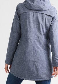 Abrigo gris con capucha, de tejido texturizado, de corte medio y puños elásticos. Presenta un yugo en la parte posterior y un dobladillo ligeramente curvado.