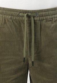 Pantaloni in velluto a coste verde oliva con una texture rigata, dotati di vita elastica con occhietti in metallo e dettagli neri.