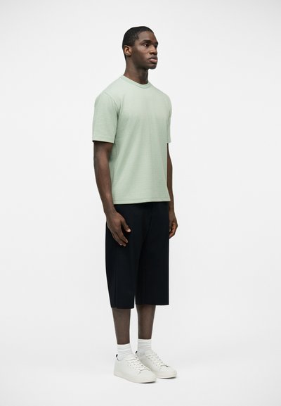 Άνδρας όρθιος, φοράει ανοιχτό πράσινο κοντομάνικο πουκάμισο, μαύρο σορτς μέχρι το γόνατο, λευκές κάλτσες και λευκά αθλητικά παπούτσια, μπροστά σε απλό λευκό φόντο.