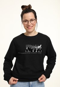 Eine lächelnde Frau in einem schwarzen Sweatshirt, das eine Grafik von sieben Männern in Anzügen zeigt. Der Text darunter zitiert einen berühmten Satz aus einem Film.