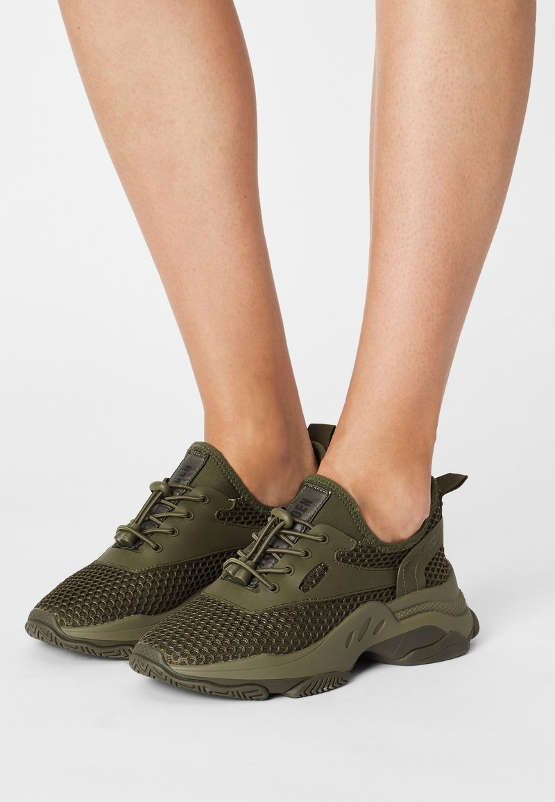 Jambes portant des baskets en mesh vert olive avec des semelles épaisses et des lacets élastiques à bascule sur fond blanc.