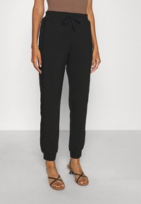 Žena v černých sportovních kalhotách s krajkovým detailem na bocích a černých sandálech na podpatku se pásky stojí před jednobarevným pozadím.