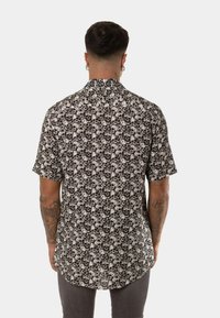 Camisa de manga corta con botones que presenta un patrón de calaveras en blanco y negro combinado con flores, confeccionada en una tela ligera, con un cuello clásico y dobladillo curvado.