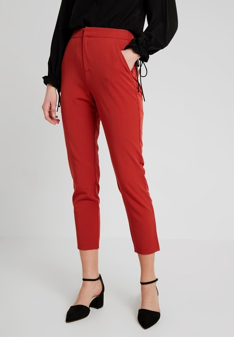 Femme portant un pantalon rouge slim cropped, un chemisier noir à manches longues et des chaussures noires à talons blocs avec bride à la cheville, debout avec une main dans la poche.